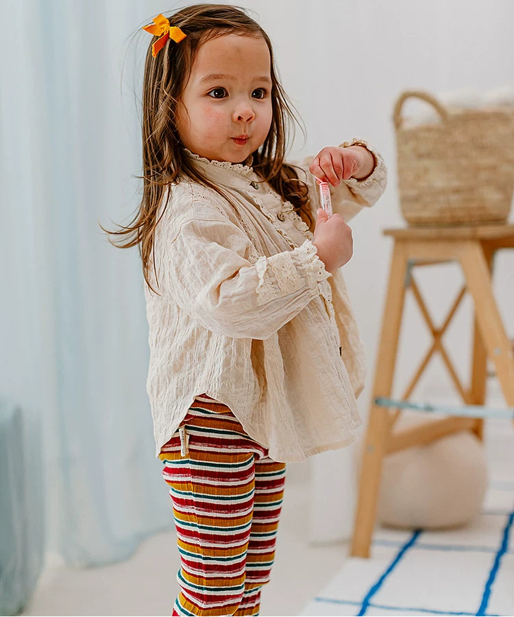A toddler girl with a yellow bow in her hair stands in a bright room wearing a cream-colored textured long-sleeve top and colorful striped pants. In the background, there is a wooden stool with a woven basket and light blue curtains.