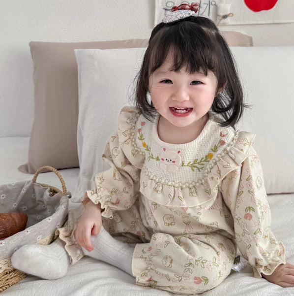 A young toddler girl with dark hair sits smiling on a white bed, wearing a cream-colored long-sleeve dress with a delicate floral print, a ruffled collar with embroidered details, and white socks. The background features neutral pillows, a decorative wall hanging, and a small woven basket.